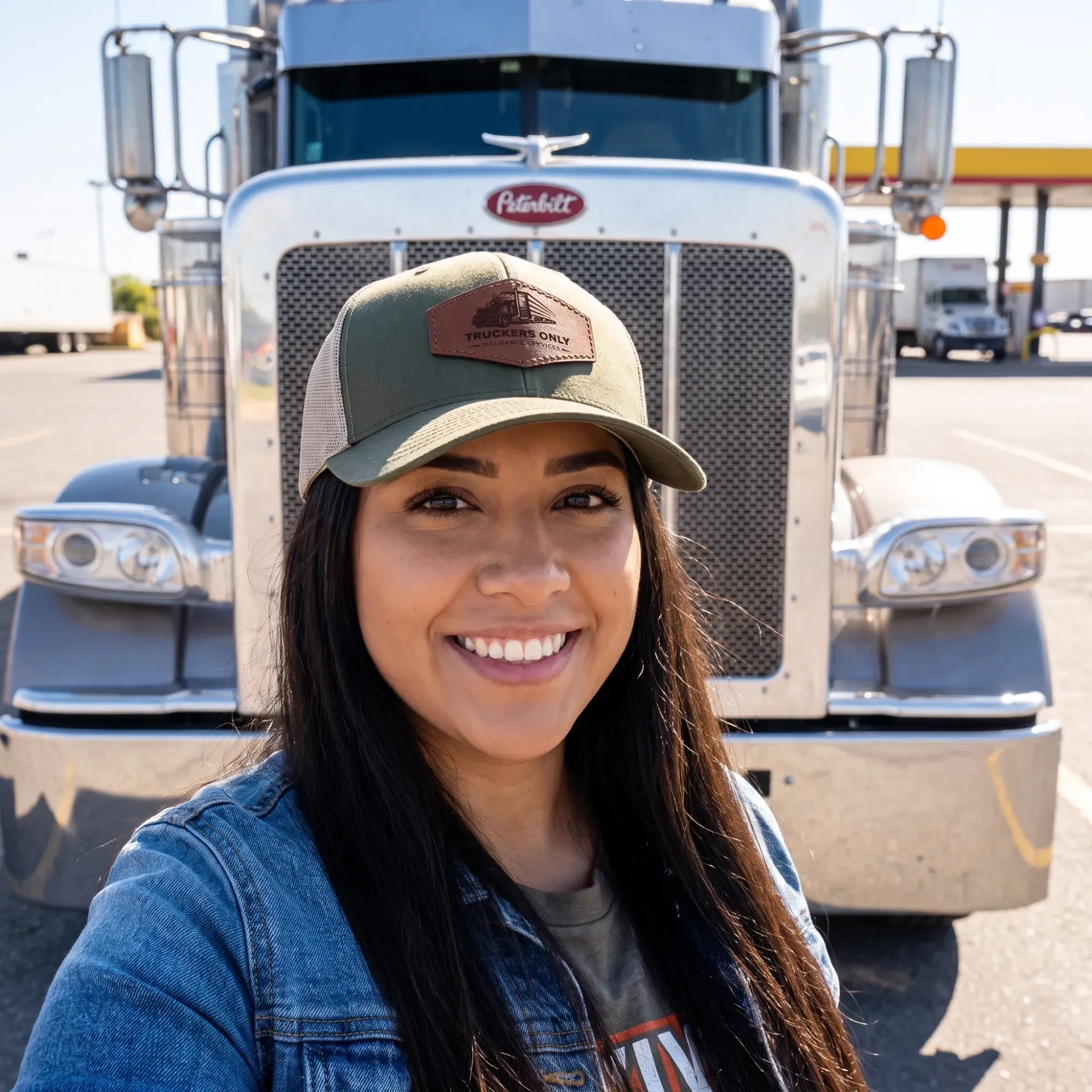 A driver wearing the 2026 Truckers Only cap in front of her Peterbilt at a fuel island