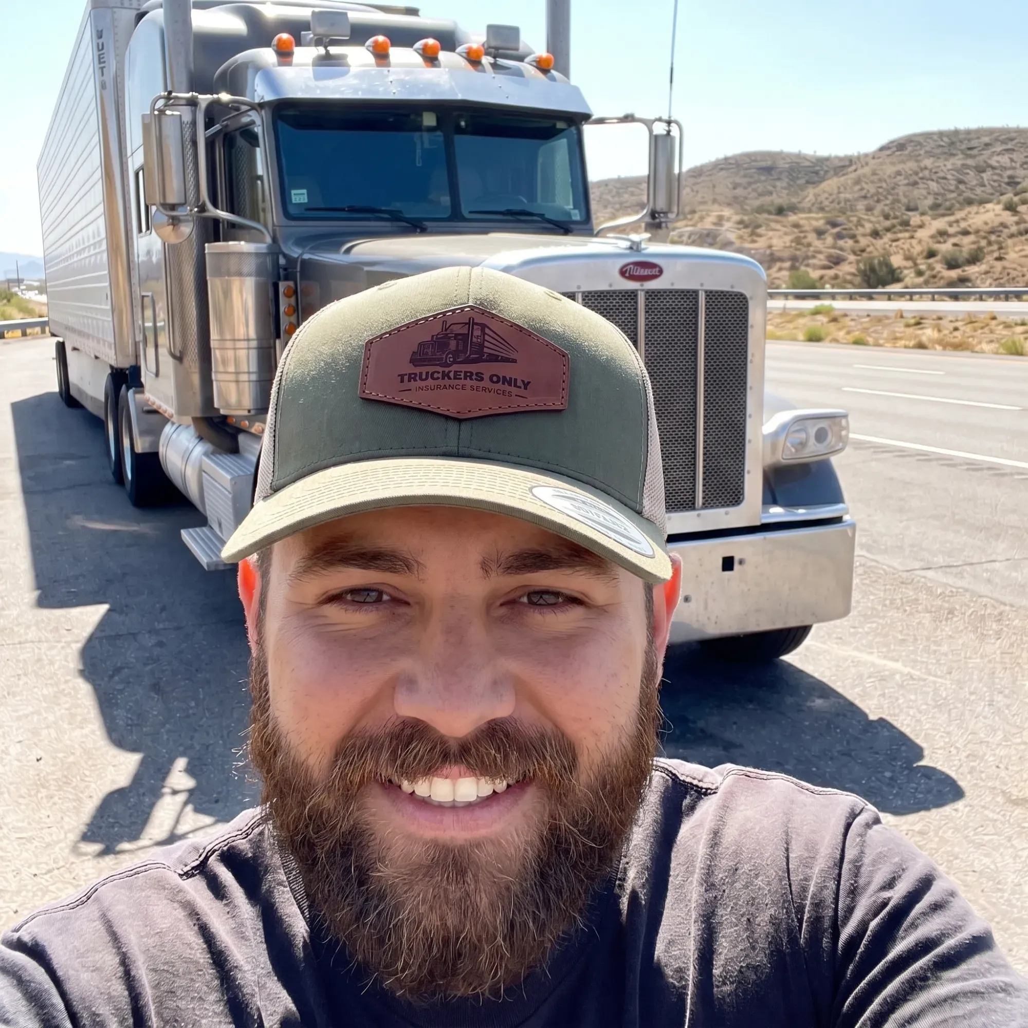 A bearded driver smiling in front of his Peterbilt on a desert highway, wearing the 2026 Truckers Only cap